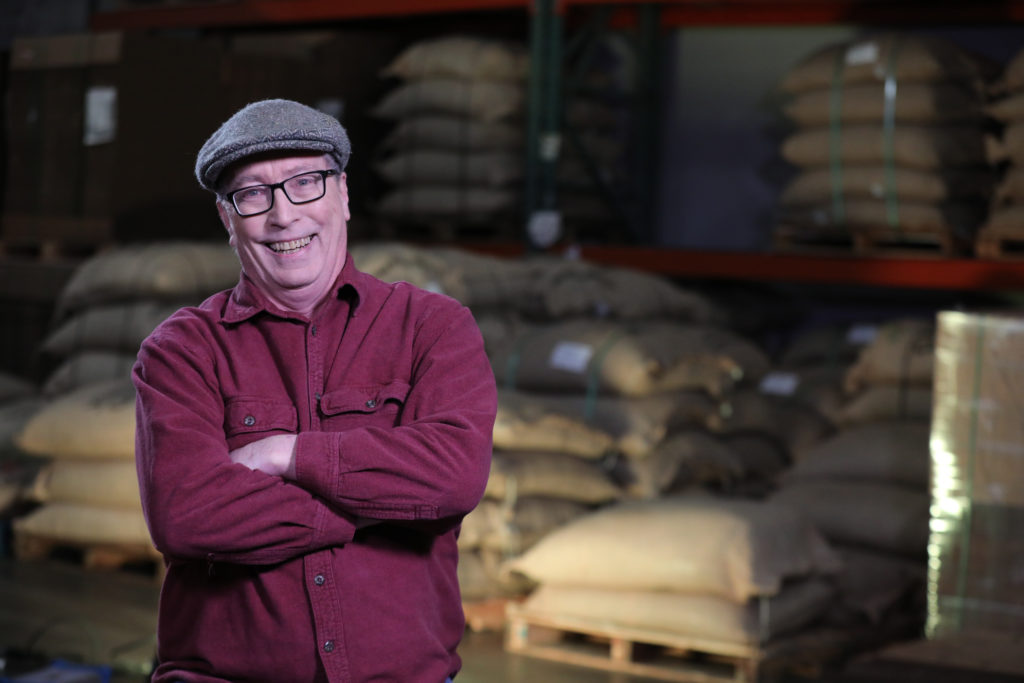 A white male-presenting person in a hat and glasses wears a red shirt and smiles at the camera. Their arms are crossed. Behind him are bags of ingredients.