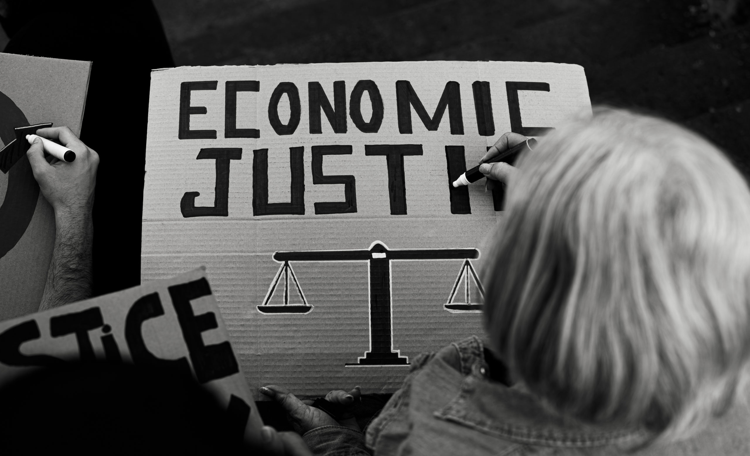 A black and white image of a person writing a protest sign that reads “Economic Justice” with an image of a justice scale.