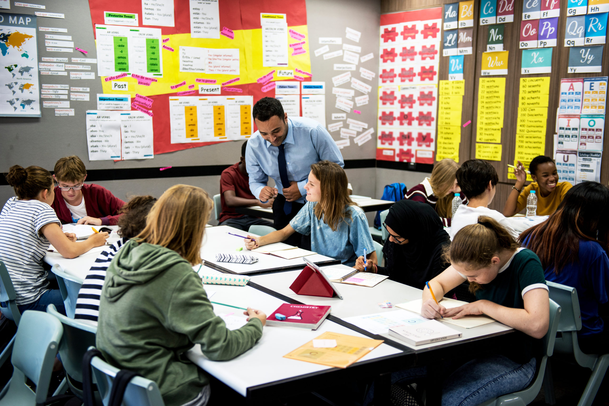 A white male-presenting middle school teacher leans over a cluster of desks as students work together on schoolwork.