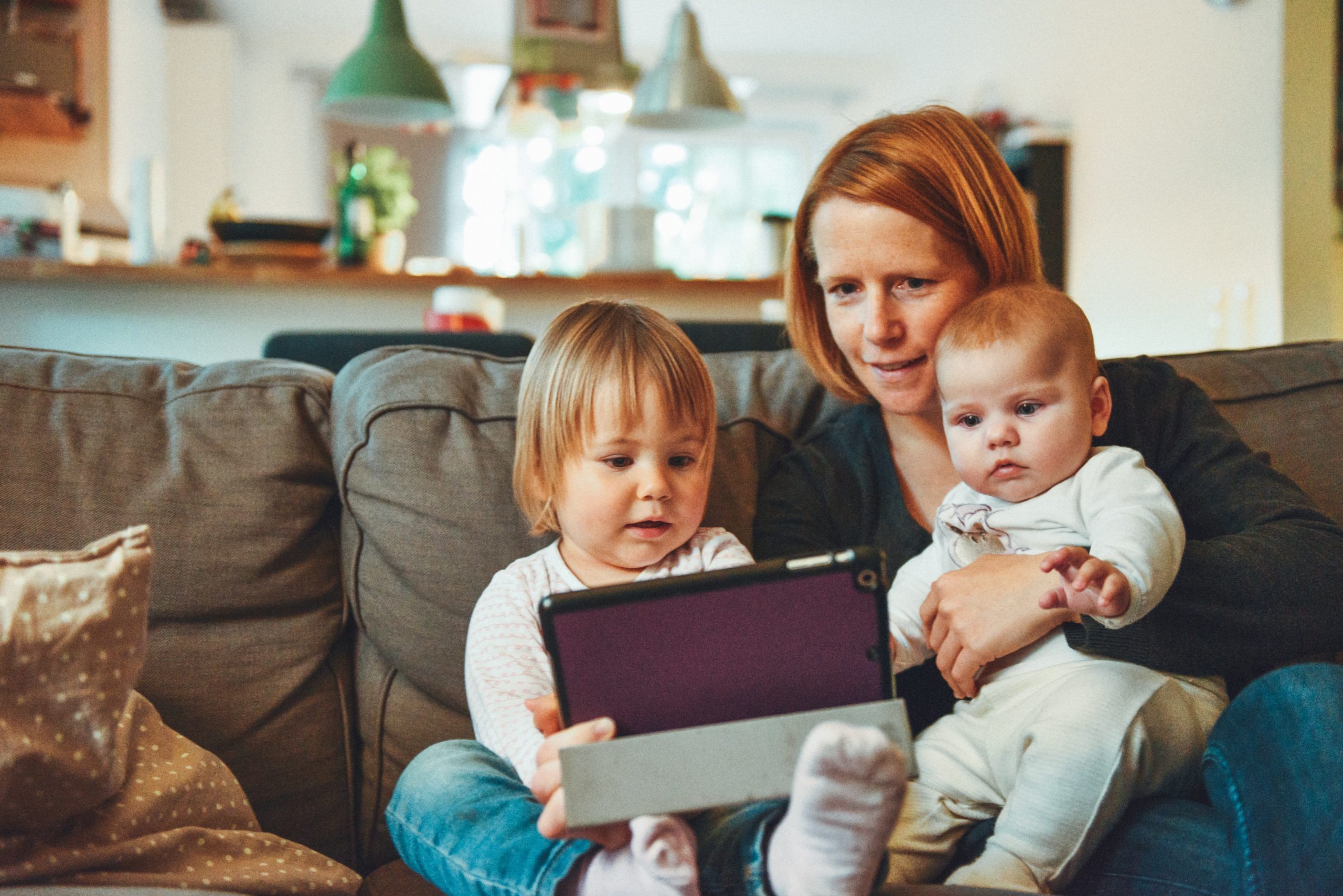 A white, female presenting person sits on a couch in her living room & reads from her tablet to her two children on her lap.