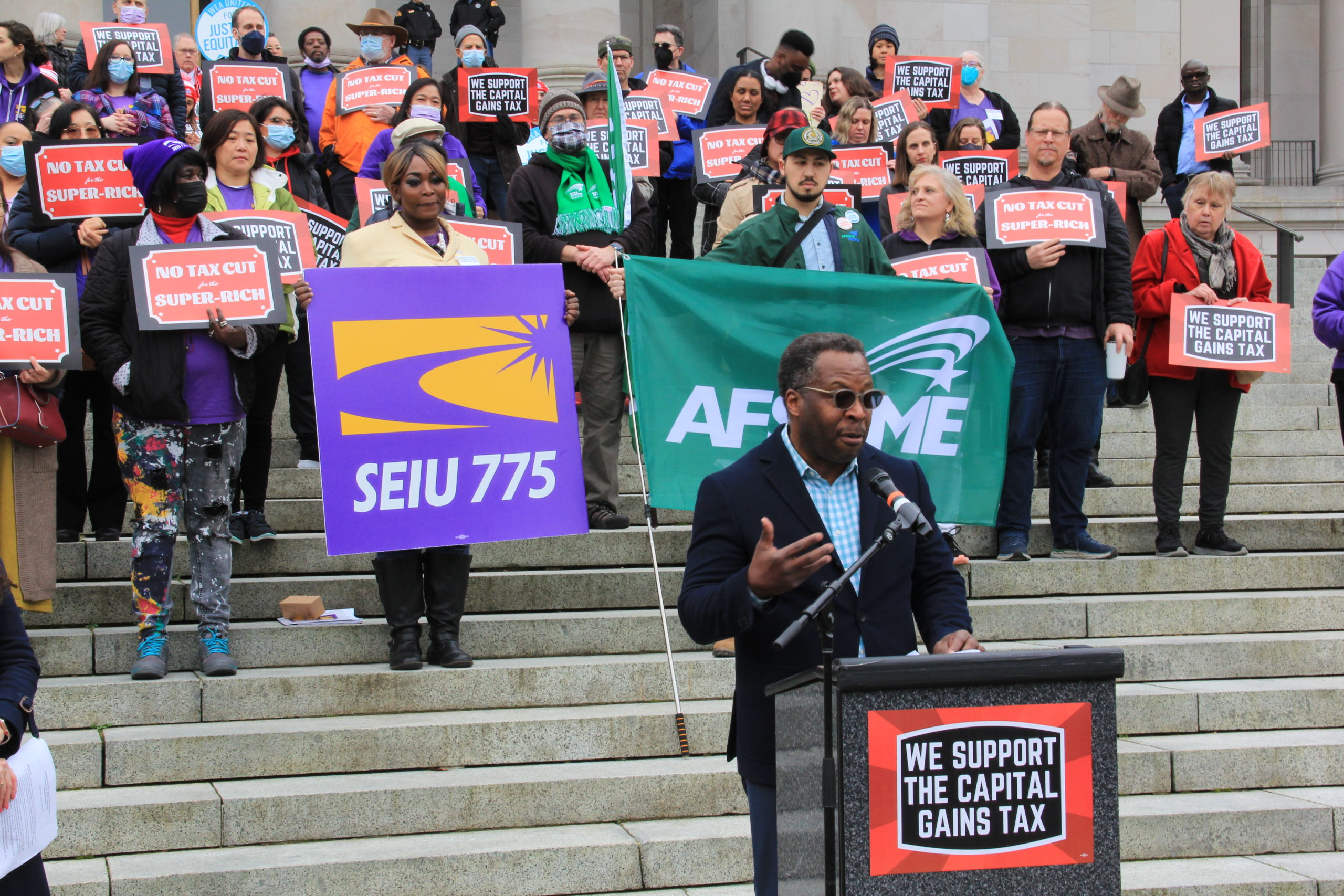Dr. Stephan Blanford, a Black man, speaks at a podium during a capital gains tax rally. Behind him are advocates from SEIU 775 and AFSCME.