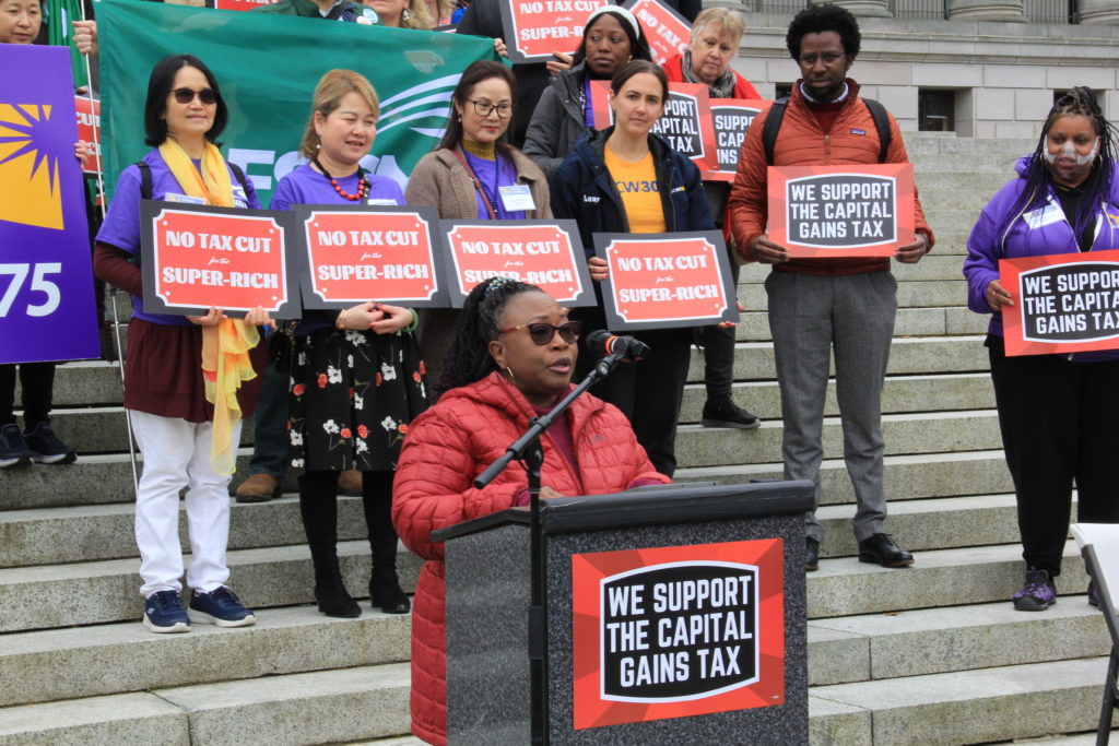 Mary Curry, a Black woman, speaks at a podium during a capital gains tax rally. SEIU 775 and AFSCME advocates are behind her.