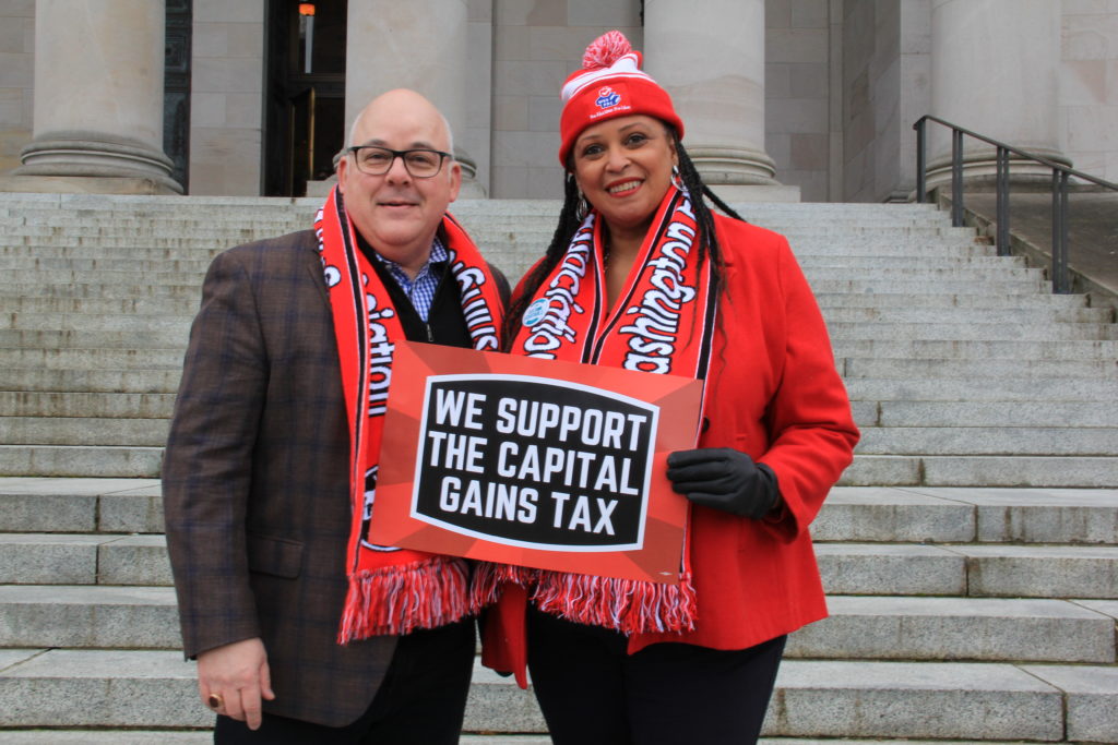 Larry Delaney and an advocate stand outside in Olympia and hold a sign that reads “We Support The Capital Gains Tax.”