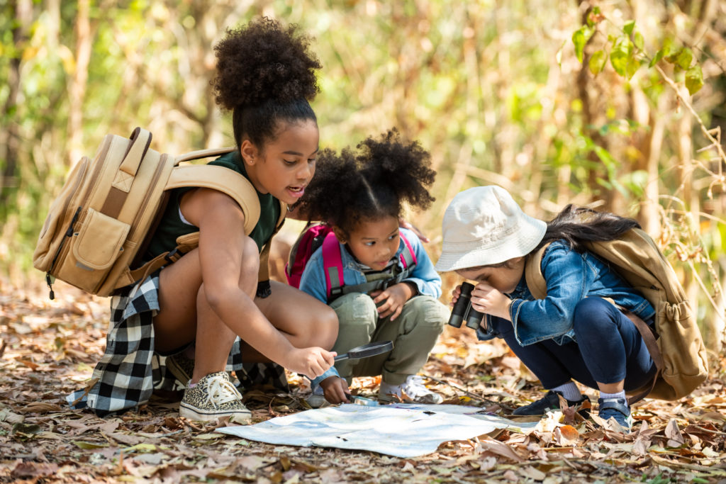 Three young kids crouch in the woods around a scavenger hunt map. One looks at the map with binoculars.