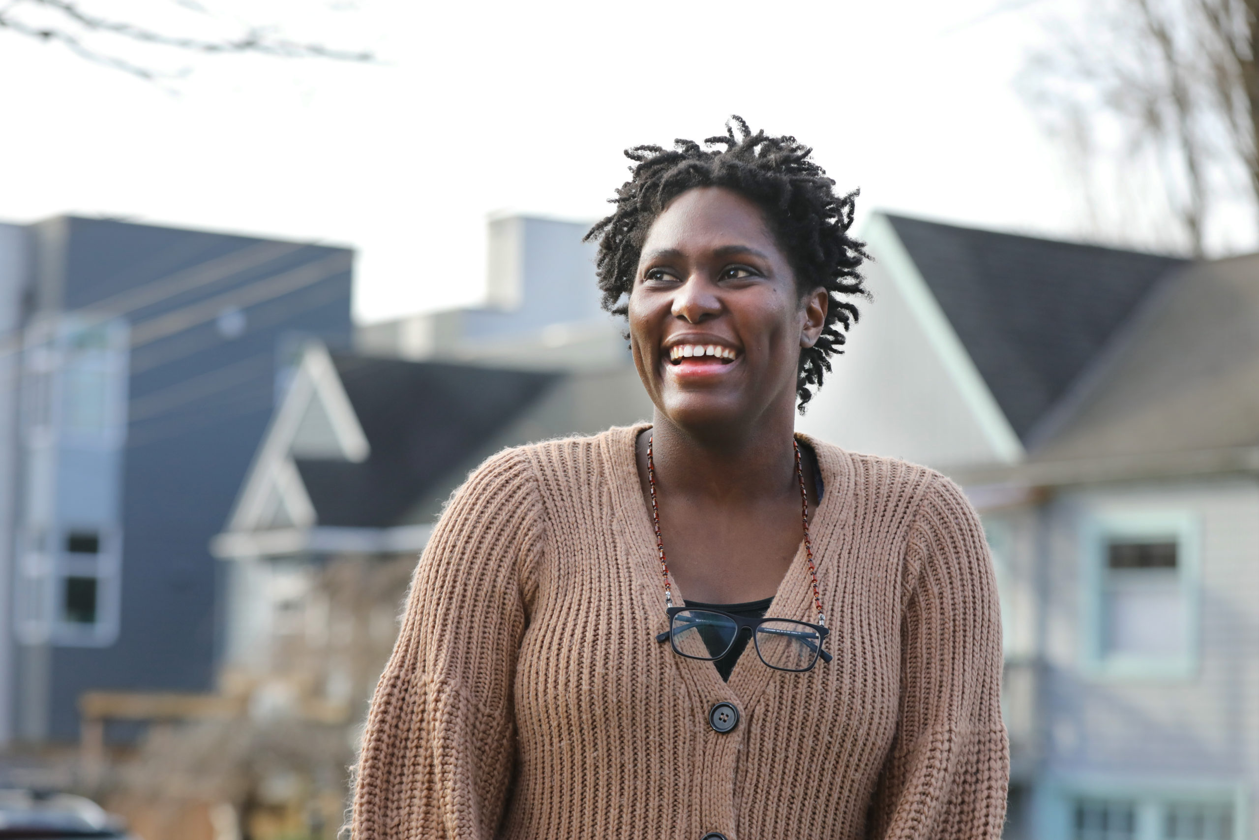 A Black female-presenting person smiles outside of their house. They are wearing glasses around their neck.
