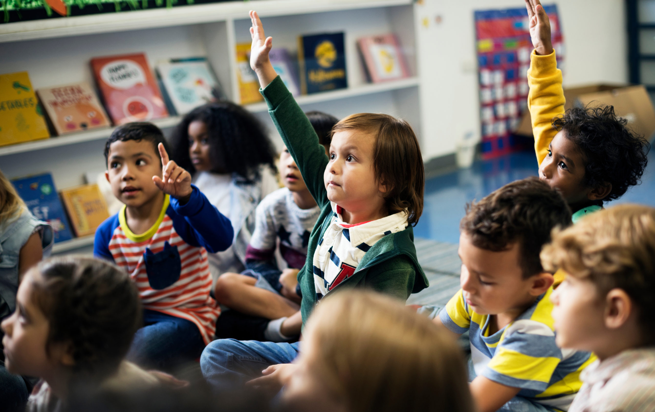 A group of kindergarten students sit on a classroom floor with books around them. Three of them raise their hands.