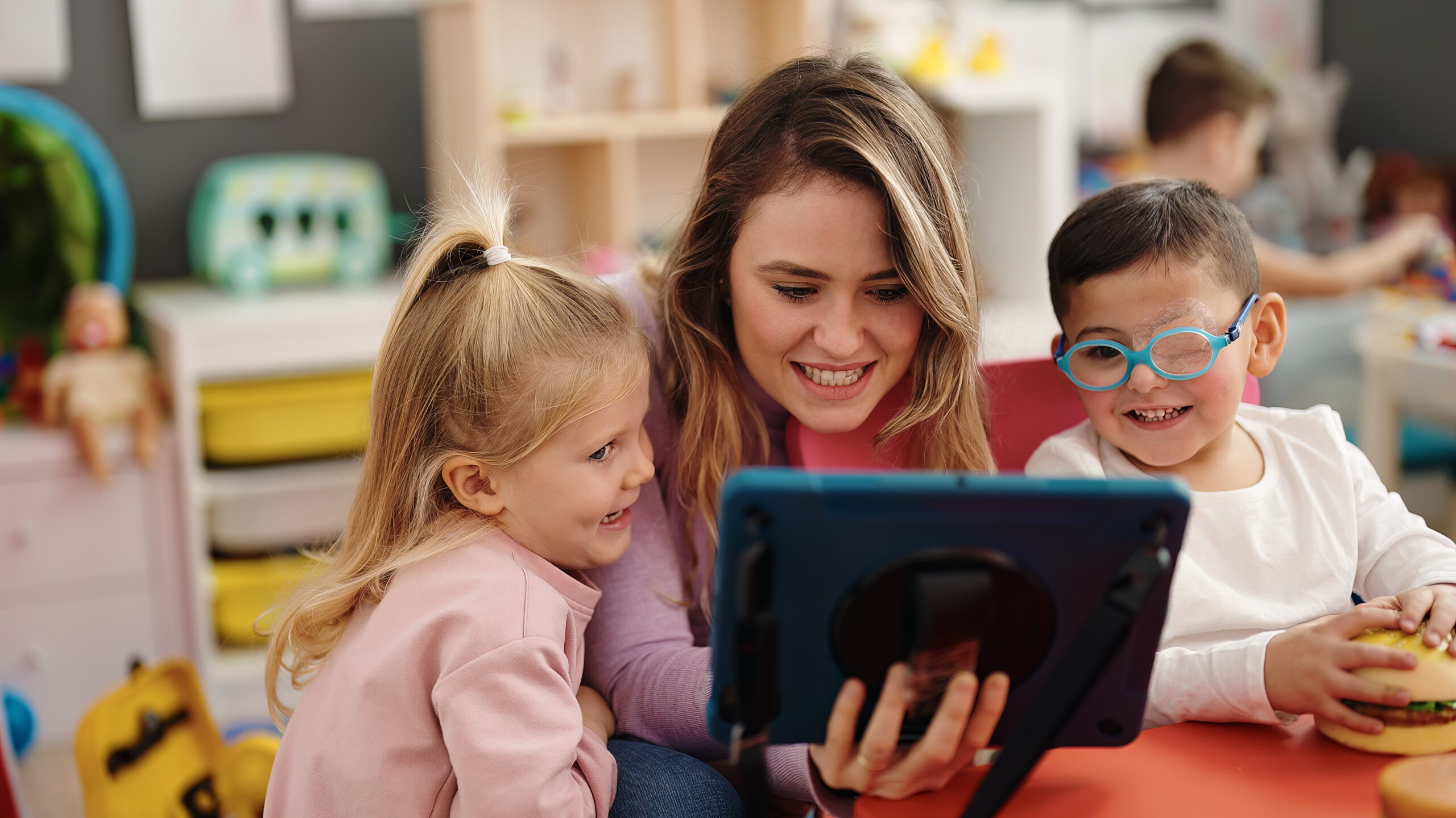 A white woman with long brown hair sits between two pre-school aged children looking at a tablet. They are in an early childhood classroom. The child on the left has blue glasses and a visual disability, the child on the left has blond hair and a pink shirt.